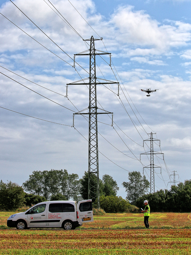 Foto von zwei Arbeitern vor einem Freileitungsmasten, der von einer autonomen Drohne inspiziert wird